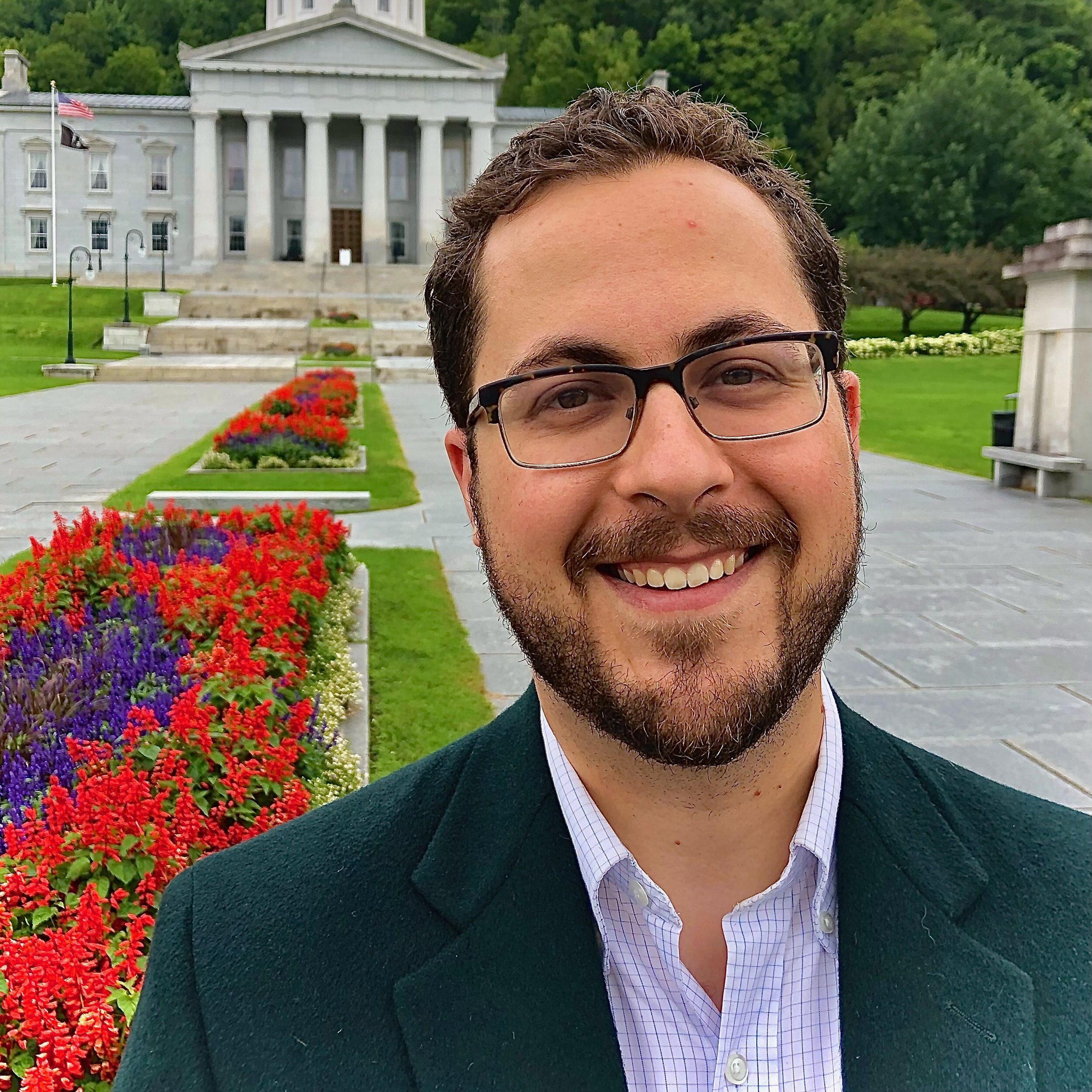 Photo of attorney Andrew Marchev. Light-skinned main with short, brown hair and a short, brown beard and mustache, He wears a dark green suit jacket and pale button up shirt. He is smiling at the camera and standing in front of landscaped flowers and a government building.