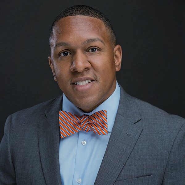 Photo of attorney Sean Fontes. Black man with very short hair, wearing a grey suit jacket, light blue buttoned shirt, and red and blue bow tie. He is smiling at the camera