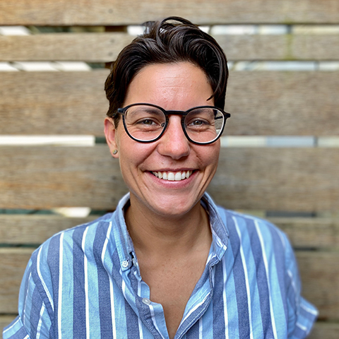 Photo of attorney Erika Dunyak. Light-skinned femme person with short, brown hair and glasses. She is smiling at the camera and wearing a short-sleeved, button up shirt with vertical light and mid-blue stripes